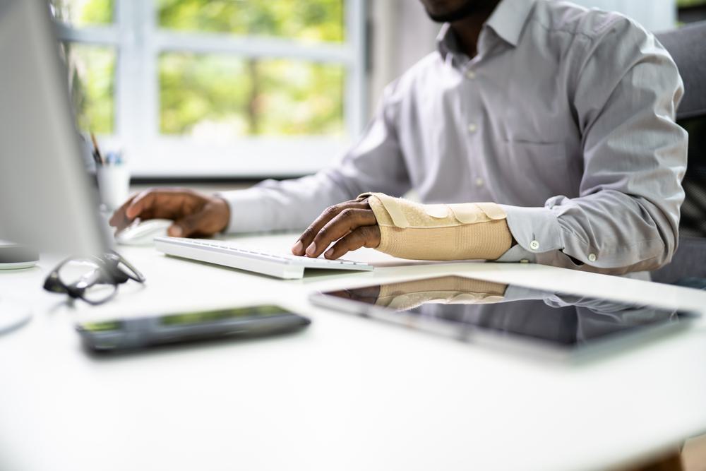 A man with a cast on his arm sits at a desk typing in front of a monitor