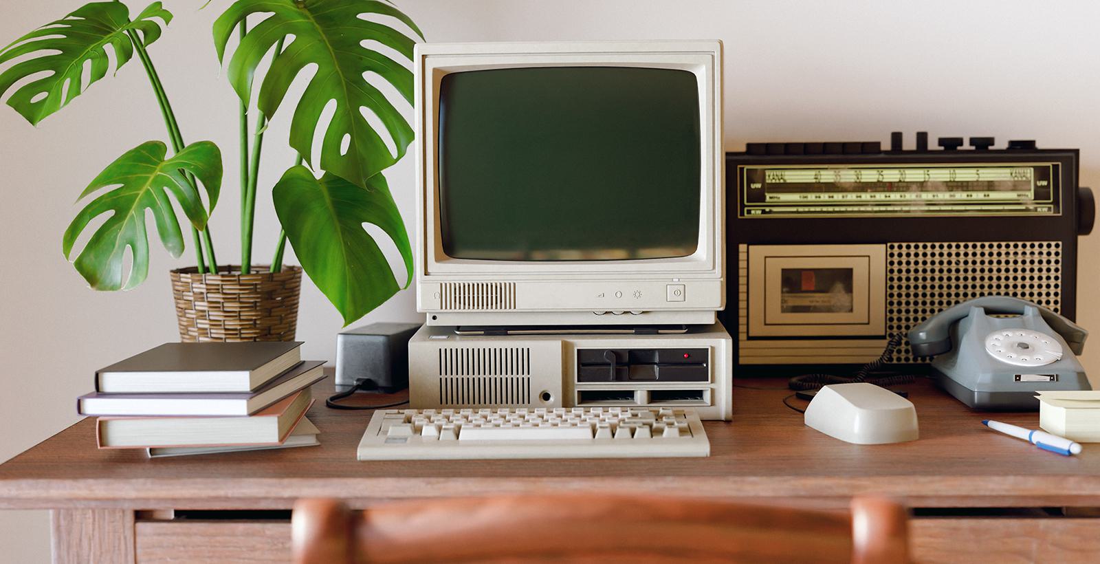 Retro computer stands on a desk with a plant, keyboard, computer mouse, retro radio, and phone.