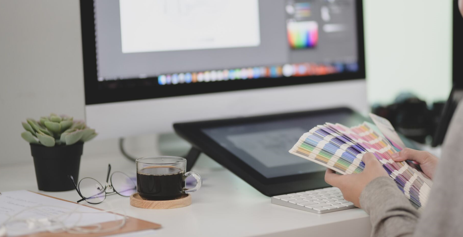 Hands sorting through colour palettes at a desk with design software on the monitor