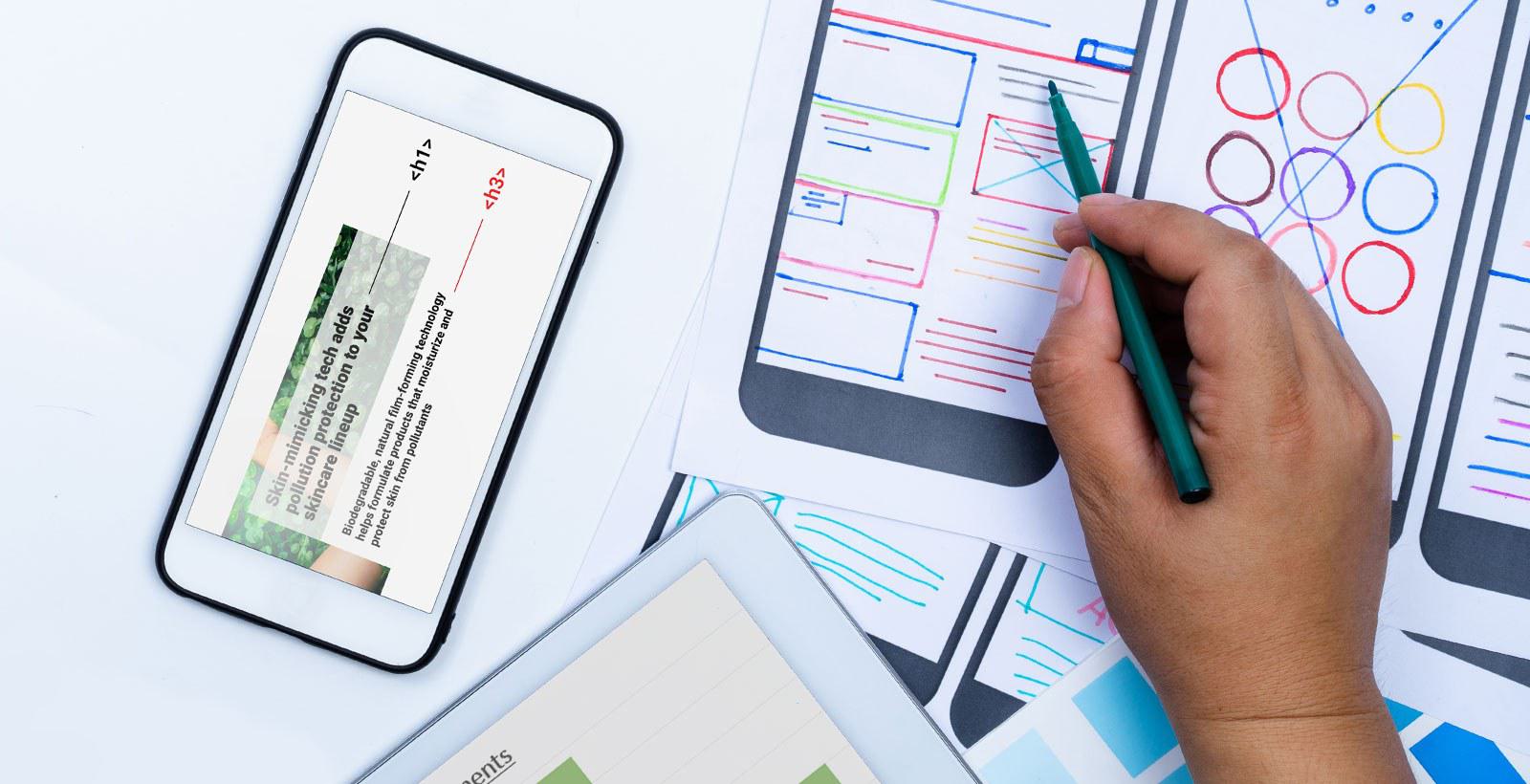 A phone displaying an accessible website next to a hand holding a pen, website designs on paper, and the corner of a tablet