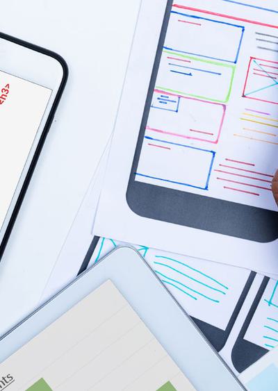 A phone displaying an accessible website next to a hand holding a pen, website designs on paper, and the corner of a tablet